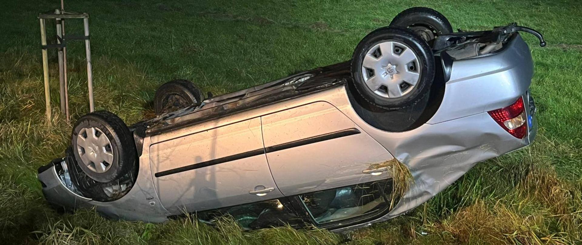 Zdjęcie zrobione nocą. Poza drogą leży na dachu samochód osobowy, oświetlany przez światła latarek.