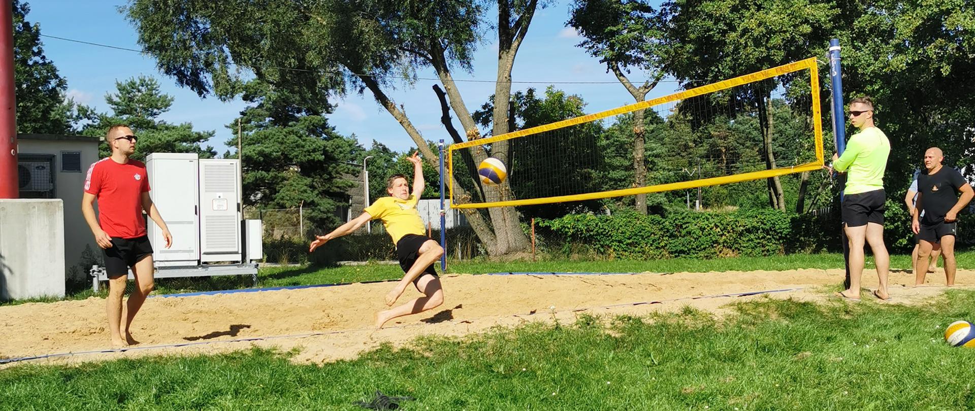 Zdjęcie przedstawia boisko do siatkówki plażowej z zawodnikami . w Otoczeniu drzewa i tereny trawiaste.
