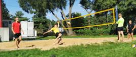 Zdjęcie przedstawia boisko do siatkówki plażowej z zawodnikami . w Otoczeniu drzewa i tereny trawiaste.