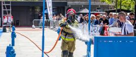 Zdjęcie przedstawia strażaka - uczestnika zawodów w pełnym umundurowaniu specjalnym wraz z aparatem ODO podającego prąd wody na tarczę. Na zdjęciu ujęto także widzów oraz sędzię zawodów.