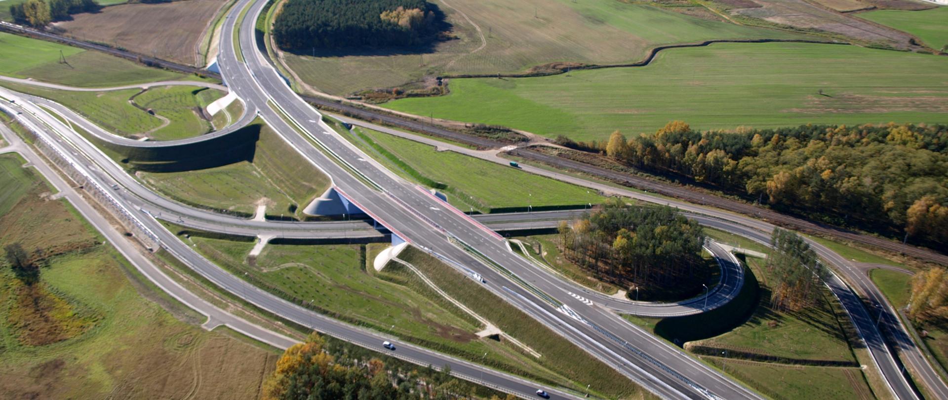 Zdjęcie przedstawiające węzeł drogowy Poznań Północ na drodze ekspresowej S11. Węzeł widoczny z powietrza przebiegający między łąkami, polami i zabudową magazynową. Na jednej z łącznic doprowadzających ruch do węzła widoczne poruszające się pojazdy.