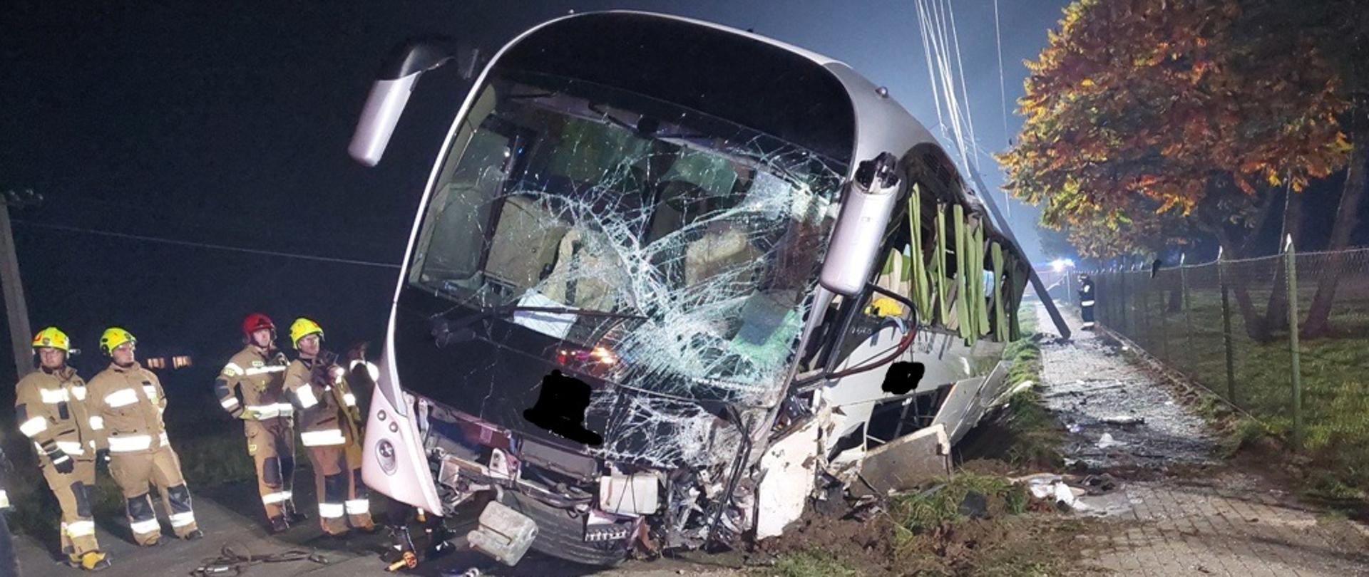 Kolorowa fotografia wykonana w nocy, przy mocnym oświetleniu sztucznym. Przedstawia widok przodu, mocno uszkodzonego, z wybitymi szybami, białego autobusu. Przy nim na drodze stoi kilku strażaków w ubraniach bojowych z hełmami na głowach. Nad autobusem widoczne przewody linii energetycznych. Z tyłu jasny blask oświetlenia terenu akcji.