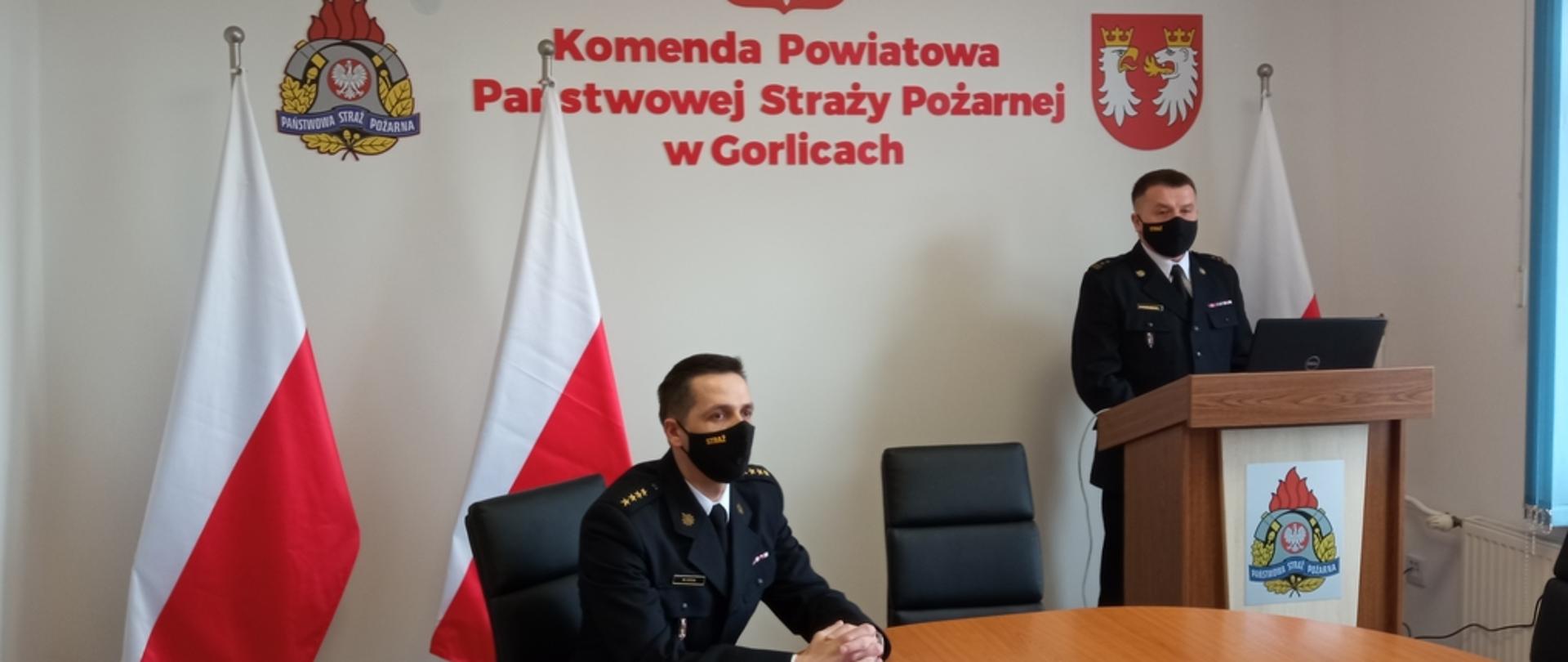 Na zdjęciu widoczny po prawej stronie na tle białej ściany z godłem Rzeczpospolitej Polskiej napisem w kolorze czerwonym Komenda Powiatowa Państwowej Straży Pożarnej w Gorlicach po bokach flagi biało czerwone przemawiający komendant powiatowy Państwowej Straży Pożarnej bryg. Krzysztof Gładysz po lewej stronie siedzący z-ca komendanta powiatowego Państwowej Straży Pożarnej w Gorlicach st.kpt. Waldemar Krok uczestniczący w rocznej odprawie. 