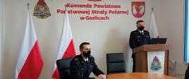 Na zdjęciu widoczny po prawej stronie na tle białej ściany z godłem Rzeczpospolitej Polskiej napisem w kolorze czerwonym Komenda Powiatowa Państwowej Straży Pożarnej w Gorlicach po bokach flagi biało czerwone przemawiający komendant powiatowy Państwowej Straży Pożarnej bryg. Krzysztof Gładysz po lewej stronie siedzący z-ca komendanta powiatowego Państwowej Straży Pożarnej w Gorlicach st.kpt. Waldemar Krok uczestniczący w rocznej odprawie. 