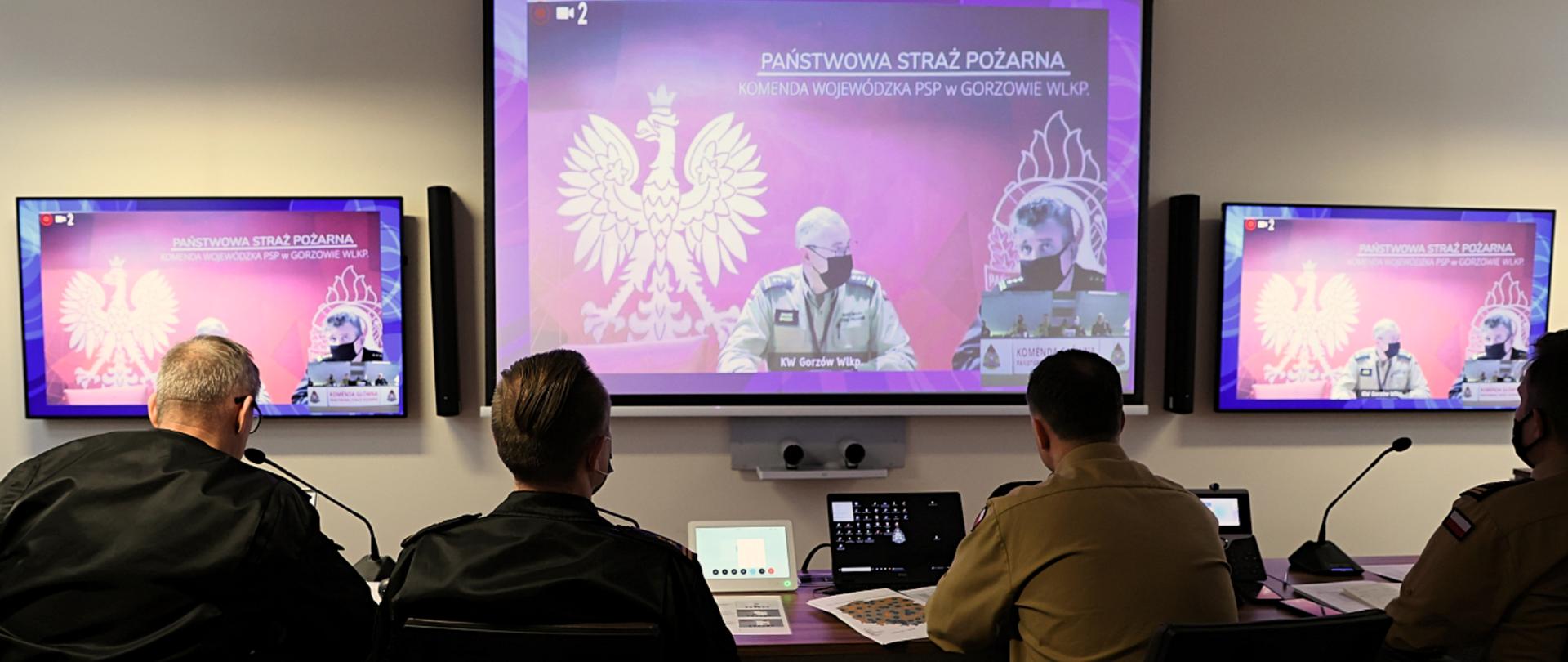 4 funkcjonariuszy siedzi przy biurku podczas wideokonferencji (widok z tyłu) , przed nimi wiszą 3 duże ekrany na których widać uczestników spotkania