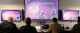 4 funkcjonariuszy siedzi przy biurku podczas wideokonferencji (widok z tyłu) , przed nimi wiszą 3 duże ekrany na których widać uczestników spotkania