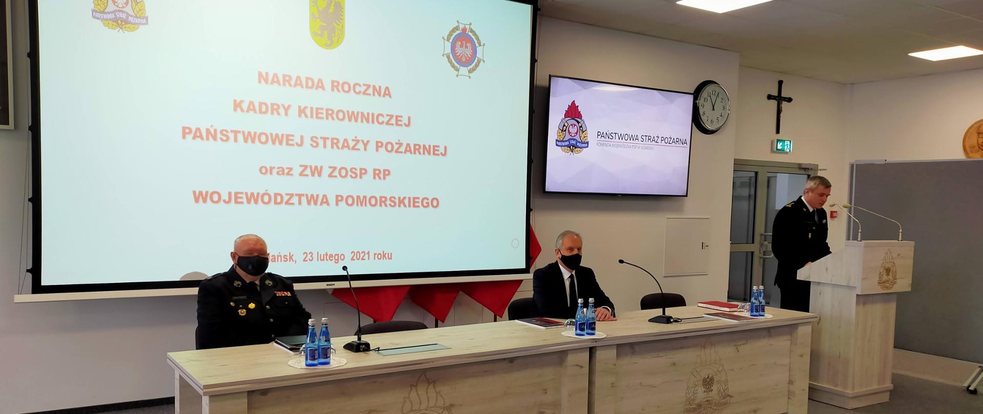 Zdjęcie przedstawia trzech mężczyzn na sali. Dwóch siedzi za stołem, z tyłu na dużym ekranie wyświetla się prezentacja. Od lewej Prezes Zarządu Wojewódzkiego OSP w mundurze, obok Wojewoda Pomorski. Po prawe stoi przy mównicy Komendant Wojewódzki PSP.