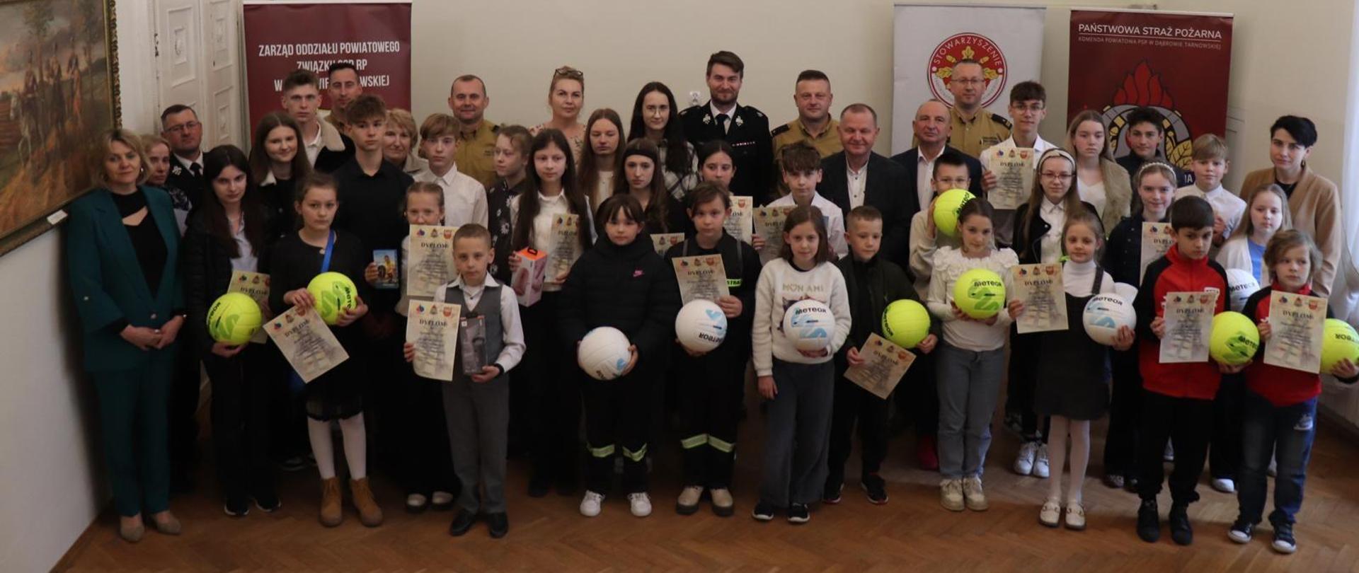 Dzieci z nagrodami, nauczyciele, opiekunowie i strażacy pozują do zdjęcia grupowego.
