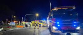 Zdjęcie 1 – na pierwszym planie widać samochód strażacki z włączonymi sygnałami świetnymi, w tle pracujący strażacy, dwie kartki pogotowia i rozbity samochód osobowy