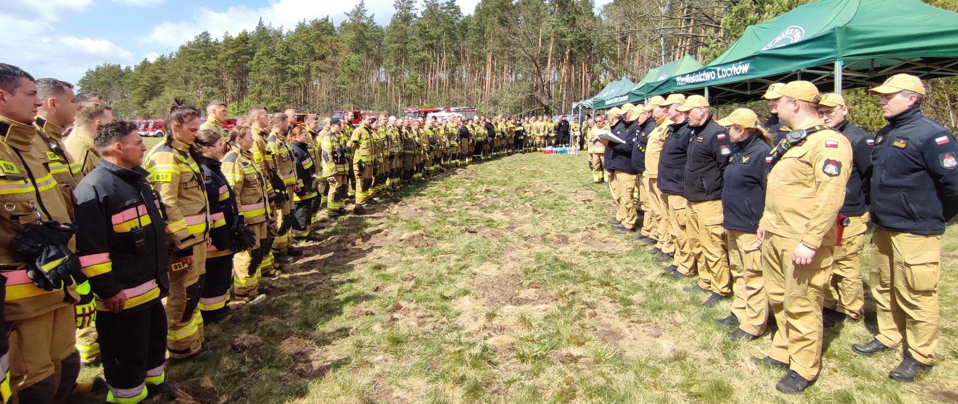 Dwie linie strażaków stoją naprzeciw siebie na trawiastym terenie, tworząc szeroki korytarz. Pośrodku znajduje się przestrzeń, w której może odbywać się prezentacja lub przemówienie. W tle widoczne są namioty i pojazdy.