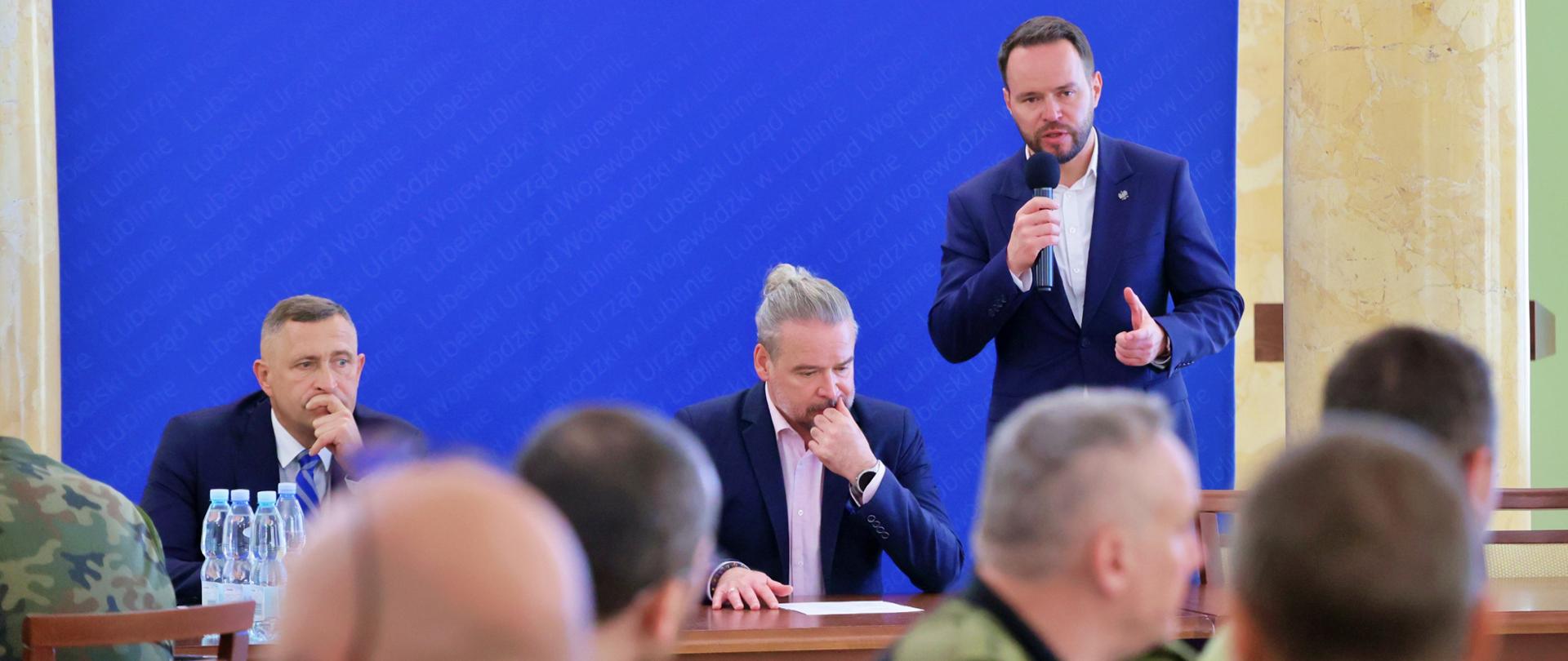 Wojewoda Lubelski Krzysztof Komorski, w granatowym garniturze, przemawia do zgromadzonych służb podczas odprawy "Bezpieczne wakacje" w Lubelskim Urzędzie Wojewódzkim. Trzymając mikrofon, gestykuluje, podkreślając wagę tematu bezpieczeństwa. W pierwszym rzędzie siedzi wicewojewoda Wojciech Wołoch (w centrum, z dłonią na brodzie) i dyrektor wydziału bezpieczeństwa i zarządzania kryzysowego LUW w Lublinie Dariusz Działo (po lewej), a także inni przedstawiciele służb mundurowych. Na ścianie za wojewodą widnieje napis "Lubelski Urząd Wojewódzki w Lublinie", potwierdzający miejsce wydarzenia.