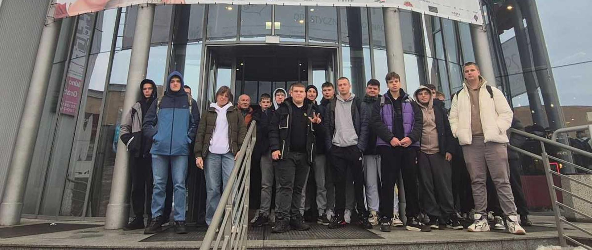 Zdjęcie przedstawia grupę młodzieży stojącej na zewnątrz przed kinem Multikino Vue we Włocławku. Za nimi znajduje się duży plakat promujący polski film familijny pod tytułem „Psoty”.
Grupa osób: Na pierwszym planie, na schodach prowadzących do wejścia, stoi kilkunastu młodych mężczyzn i chłopców, ubranych w zimowe kurtki i dżinsy. Patrzą w kierunku aparatu. Tło stanowi przeszklona fasada budynku kina.