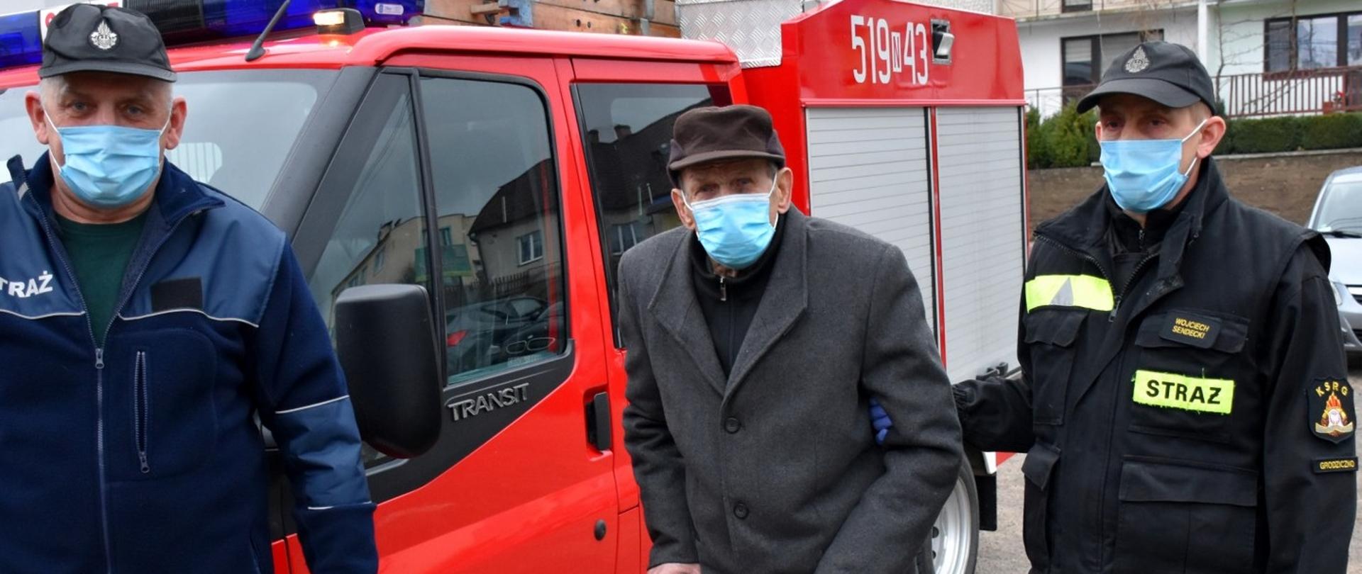 Starszy Pan z laską w towarzystwie dwóch strażaków. Wszyscy zgodnie z obowiązującymi przepisami posiadają maseczki ochronne na twarzy. 