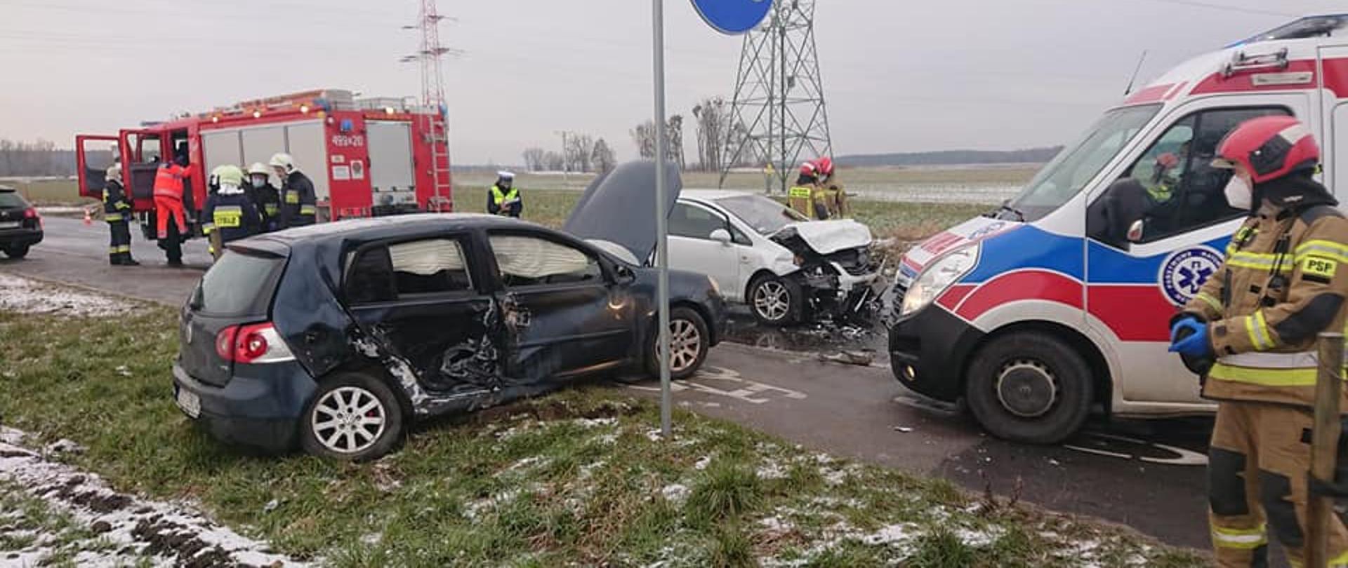Zdjęcie przedstawia dwa rozbite samochody osobowe po wypadku oraz służby ratownicze w tym strażaków prowadzących działania ratownicze. Na kadrze widoczny również samochód pożarniczy i ambulans zespołu ratownictwa medycznego.