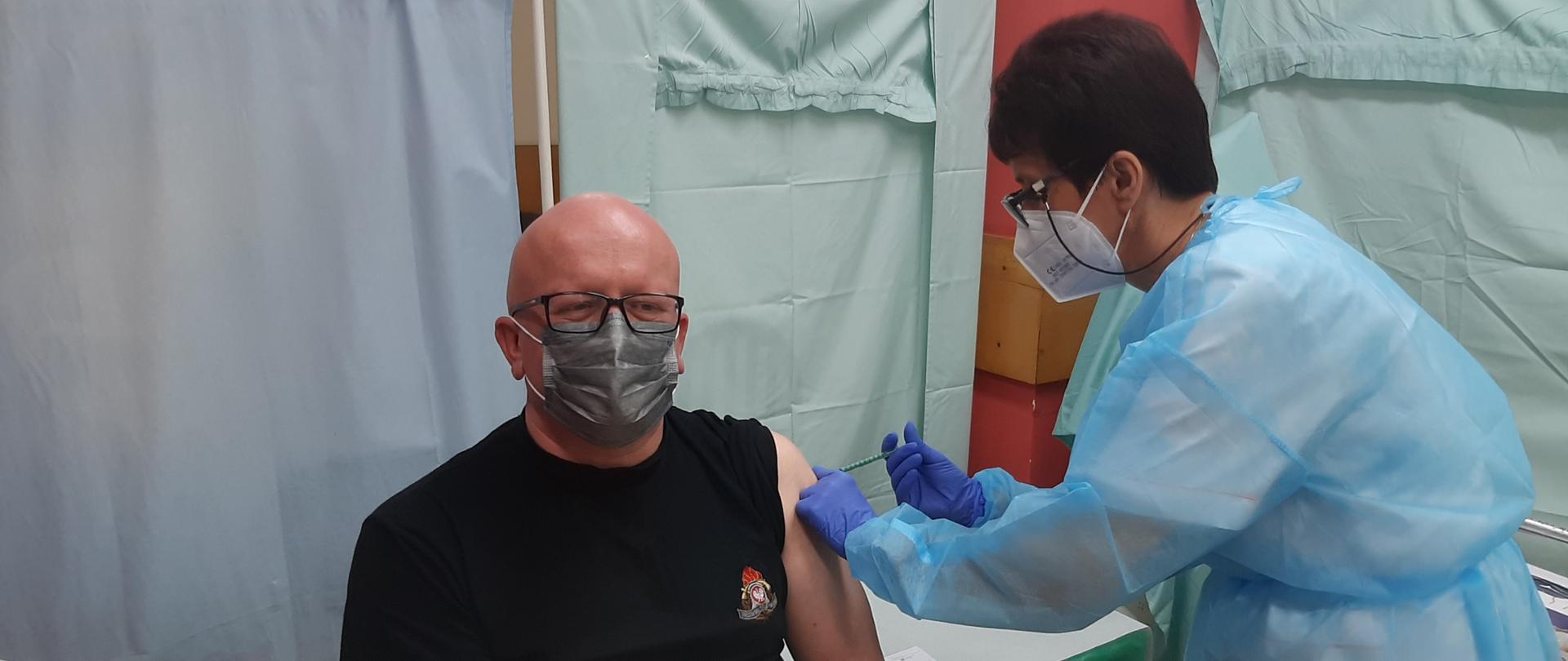 Zdjęcie przedstawia mł.bryg. Cezarego Wochnę z komendy Powiatowej PSP w Koluszkach w czasie szczepienia przeciwko COVID-19, z prawej strony pielęgniarka wykonująca zastrzyk . 