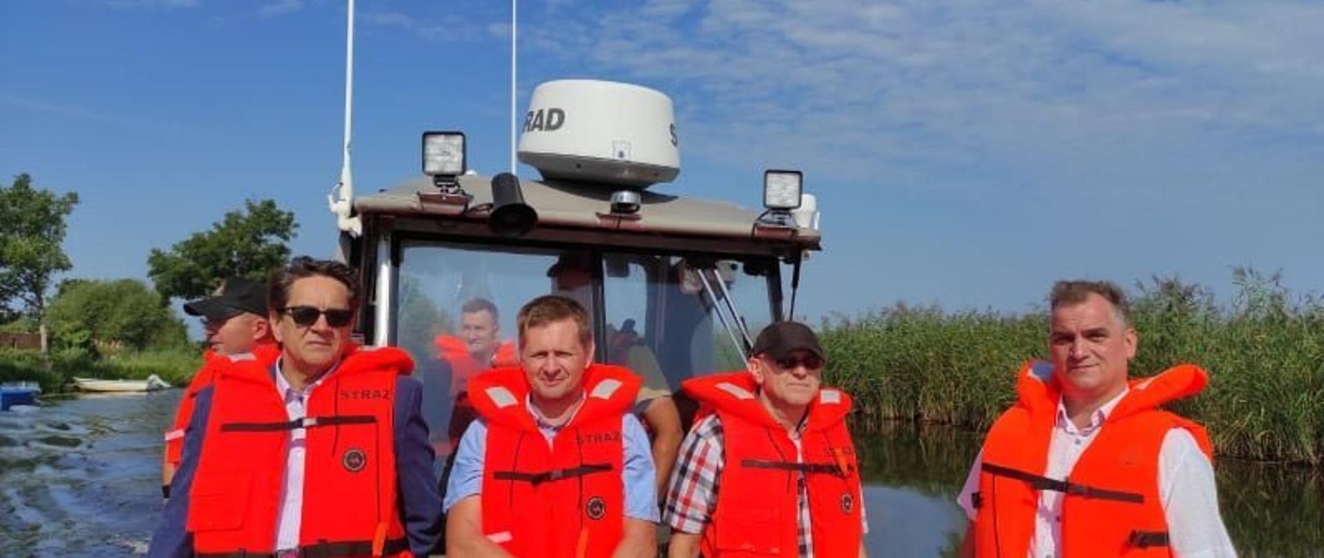 Na zdjęciu widoczna kabina łodzi. Na łodzi samorządowcy oraz przedstawiciele służb w pomarańczowych kamizelkach. Słonecznie, niebieskie niebo, z prawej strony zielone trzciny.