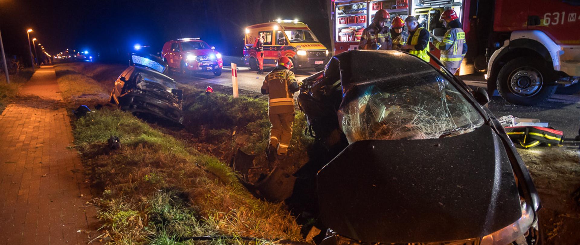 Na zdjęciu widoczne dwa rozbite samochody koloru ciemnego, stojące w przydrożnym rowie. Na jezdni stoją samochody strażackie koloru czerwonego i żółty ambulans. Przy samochodach widoczni strażacy w czerwonych hełmach i policjant w odblaskowej, żółtej kamizelce. Zdjęcie wykonane w porze nocnej.