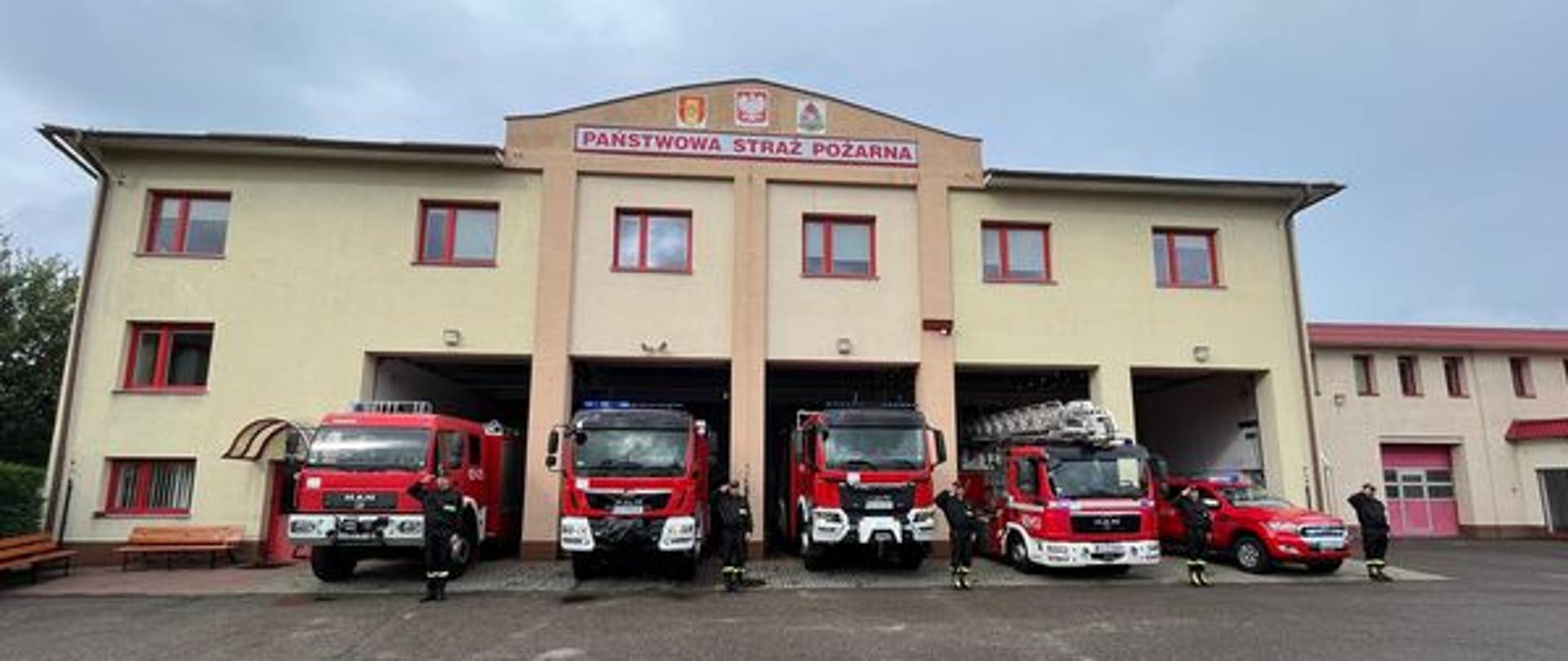 Zdjęcie przedstawia budynek jednostki ratowniczo-gaśniczej w Człuchowie, w otwartych bramach stoją samochody pożarnicze z włączonymi sygnałami świetlnymi i dźwiękowymi. Przy samochodach stoją strażacy oddający honory. 