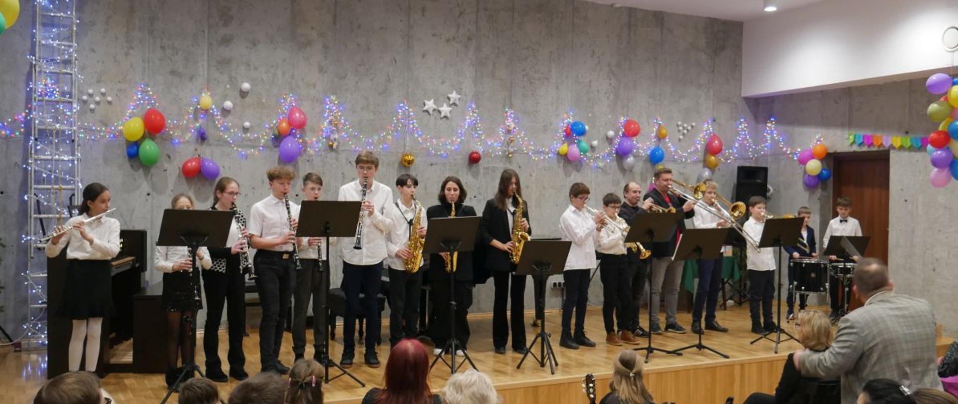 Zdjęcie przedstawia grupę uczniów - orkiestrę dętą - na scenie szkoły muzycznej. Na pierwszym planie siedząca twarzą do sceny publiczność, scena udekorowana jest balonami i lampkami choinkowymi.