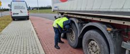 Na pierwszym planie (od prawej): umundurowany inspektor opolskiej Inspekcji Transportu Drogowego kontroluje stan techniczny zatrzymanej ciężarówki - stoi obok naczepy i świeci latarką na tarcze hamulcowe przy kole. W tle: tył oznakowanego furgonu opolskiej Inspekcji Transportu Drogowego.