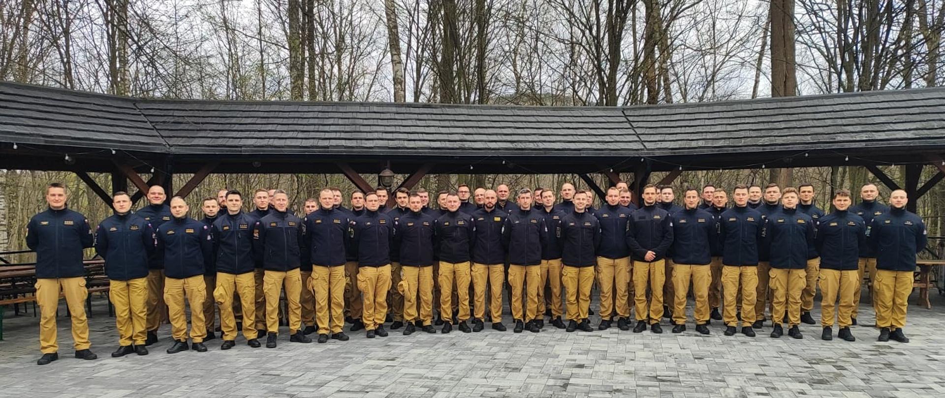 Grupa kilkudziesięciu strażaków Państwowej Straży Pożarnej stoi w szeregu na zewnątrz pod zadaszoną wiatą, ubrani w granatowe bluzy i piaskowe spodnie; w tle drzewa bez liści.