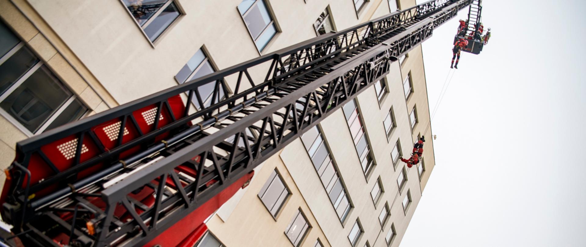 Drabina mechaniczna rozstawiona, w tle strażak zjeżdżający z kosza drabiny po linie