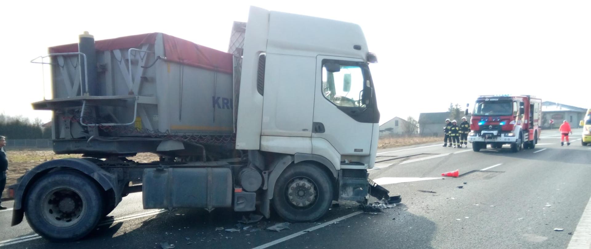 Zdjęcie przedstawia drogę krajową numer 74. Po lewej stronie stoi uszkodzony ciągnik siodłowy z naczepą. W głębi widać zaparkowany na jezdni samochód pożarniczy oraz stoi grupa strażaków. Skrajnie po prawej stoi ratownik medyczny.