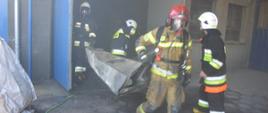 Pożar lakierni. Teren jednej z firm w miejscowości Dłoń. czterech strażaków wynosi z budynku objętego wcześniej pożarem, zniszczony przez ogień fragment urządzenia technologicznego. 