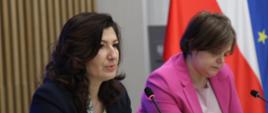 Zdjęcie z boku pod kątem, za stołem siedzi wiceminister Zioło-Pużuk w różowej marynarce i mówiąca do małego mikrofonu kobieta w czarnej marynarce, za nimi ściana z pionowych listewek, z prawej pod ścianą flagi Polski i UE. 