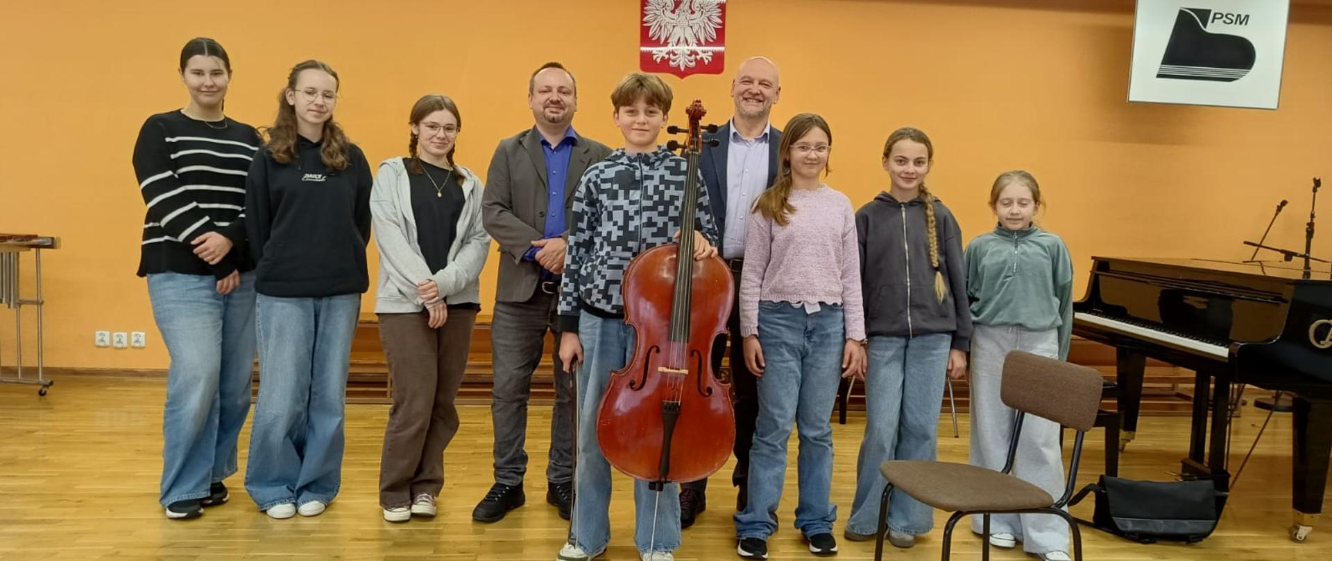 Uczniowie oraz dwóch nauczycieli stoją na scenie w rzędzie, Na środku stoi uśmiechnięty uczeń i trzyma wiolonczele. W tle żółta ściana.