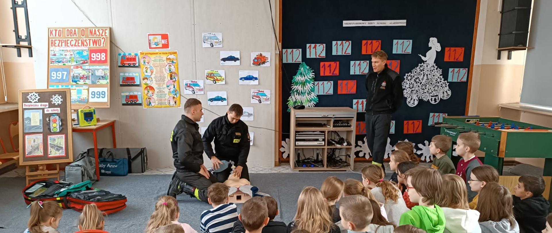 Dwóch strażaków pochylonych nad manekinem robią pokaz pierwszej pomocy. Trzeci strażak stoi przed nimi. Pokazowi przygląda sie kilkanaście małych dzieci. Zdjęcie w klasię. Na ścianach wiszą obrazki pojazdów pożarniczych oraz pogotowia i policji. Tablica wyklejona napisami z numerem 112 