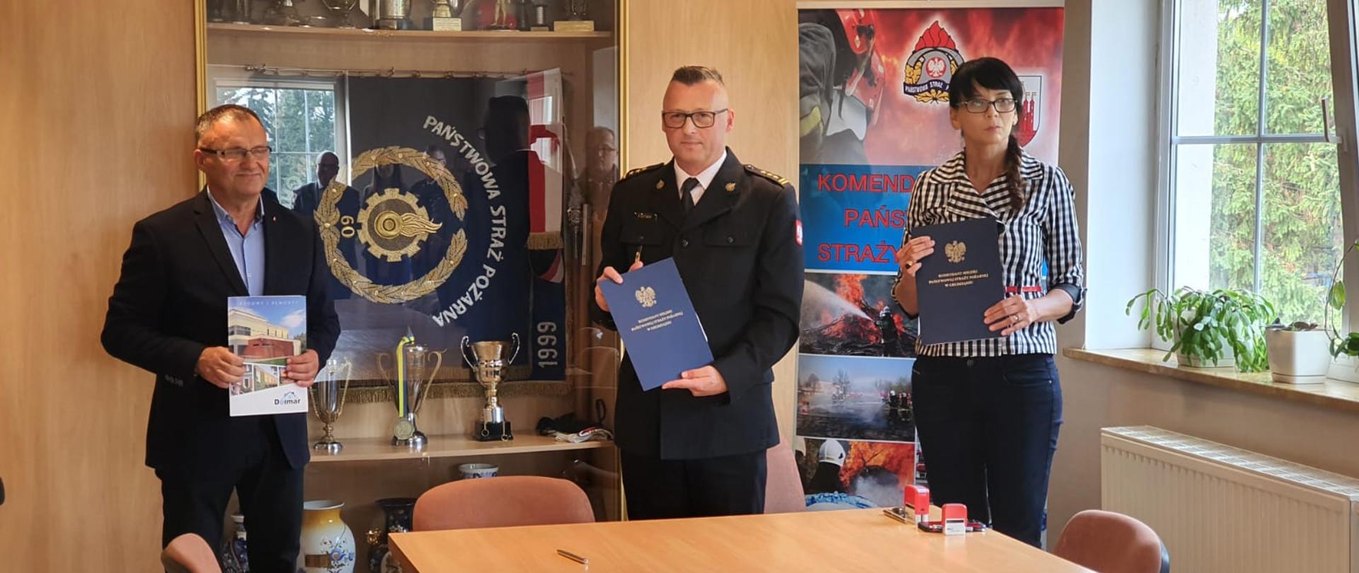 W gabinecie stoją trzy osoby - wykonawca robót budowlanych, komendant i ksiegowa. W ręku trzymają dokumenty, za nimi gablota ze sztandarwm, dalej okno.