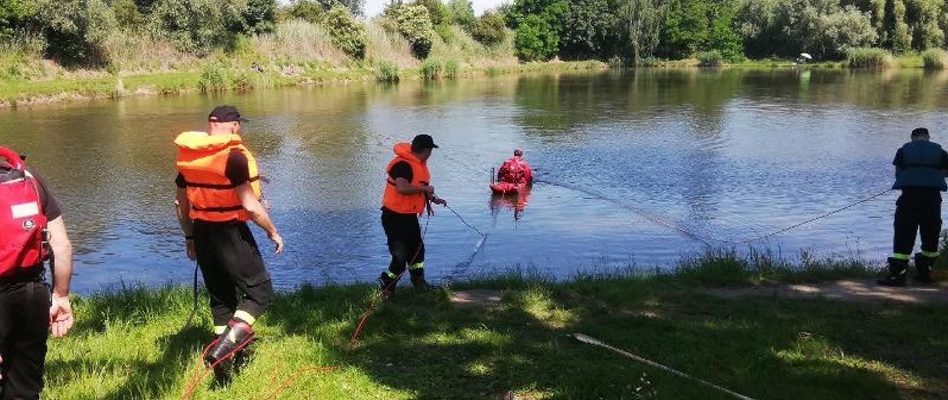 Trzech strażaków w kamizelkach ratunkowych trzyma linę asekuracyjną która podpięta jest do deski lodowej. Na desce lodowej ratownicy podejmują działania na wodzie. W tle widać tereny zielone i słoneczną pogodę.
