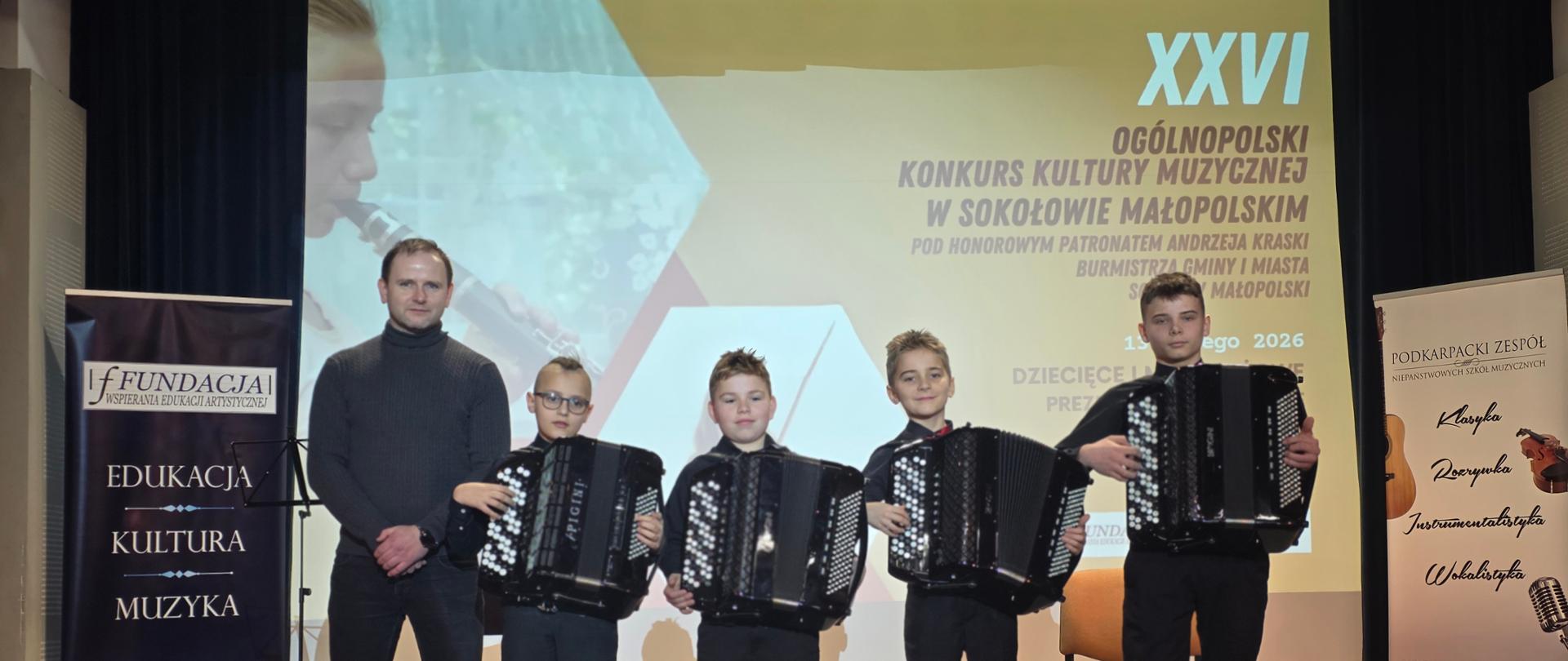 Szersze ujęcie sceny, na której czterech młodych akordeonistów stoi w rzędzie, każdy trzyma instrument. Obok nich stoi starszy mężczyzna. Tło z ekranem z grafiką konkursu jest widoczne, podobnie jak banery po bokach.