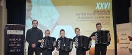 Szersze ujęcie sceny, na której czterech młodych akordeonistów stoi w rzędzie, każdy trzyma instrument. Obok nich stoi starszy mężczyzna. Tło z ekranem z grafiką konkursu jest widoczne, podobnie jak banery po bokach.