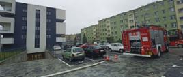 Na osiedlu bloków, strażacy wypompowują wodę z zalanego parkingu podziemnego. Po prawej ustawiony tyłem samochód pożarniczy, obok auta osobowe na parkingu. Po lewej blok z wjazdem do parkingu podziemnego. Przy otwartej bramie stoją strażacy w ubraniach specjalnych koloru pisakowego. W tle kolejne bloki osiedla. 