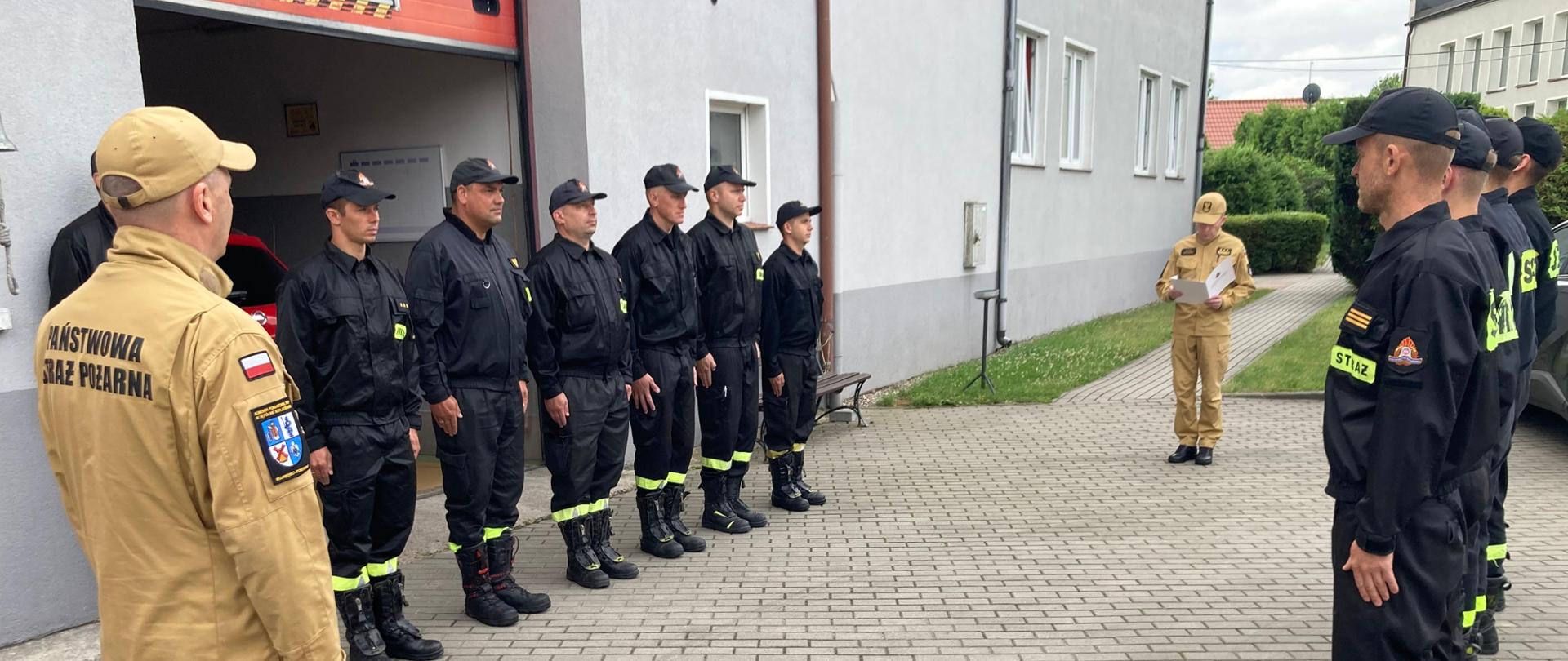 Strażacy państwowej straży pożarnej w mundurach koszarowych podczas uroczystej zmiany służbowej stoją naprzeciw siebie na baczność, między nimi dowódca jednostki Robert Liss oraz komendant powiatowy państwowej straży pożarnej starszy brygadier Maciej Suchomski.