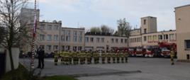 Plac przed jednostką. Poczet flagowy podnosi flagę na maszt, Ustawieni strażacy w mundurach specjalnych z dowódcą oddającym honor. W tle pojazdy JRG Tczew i budynek komendy.