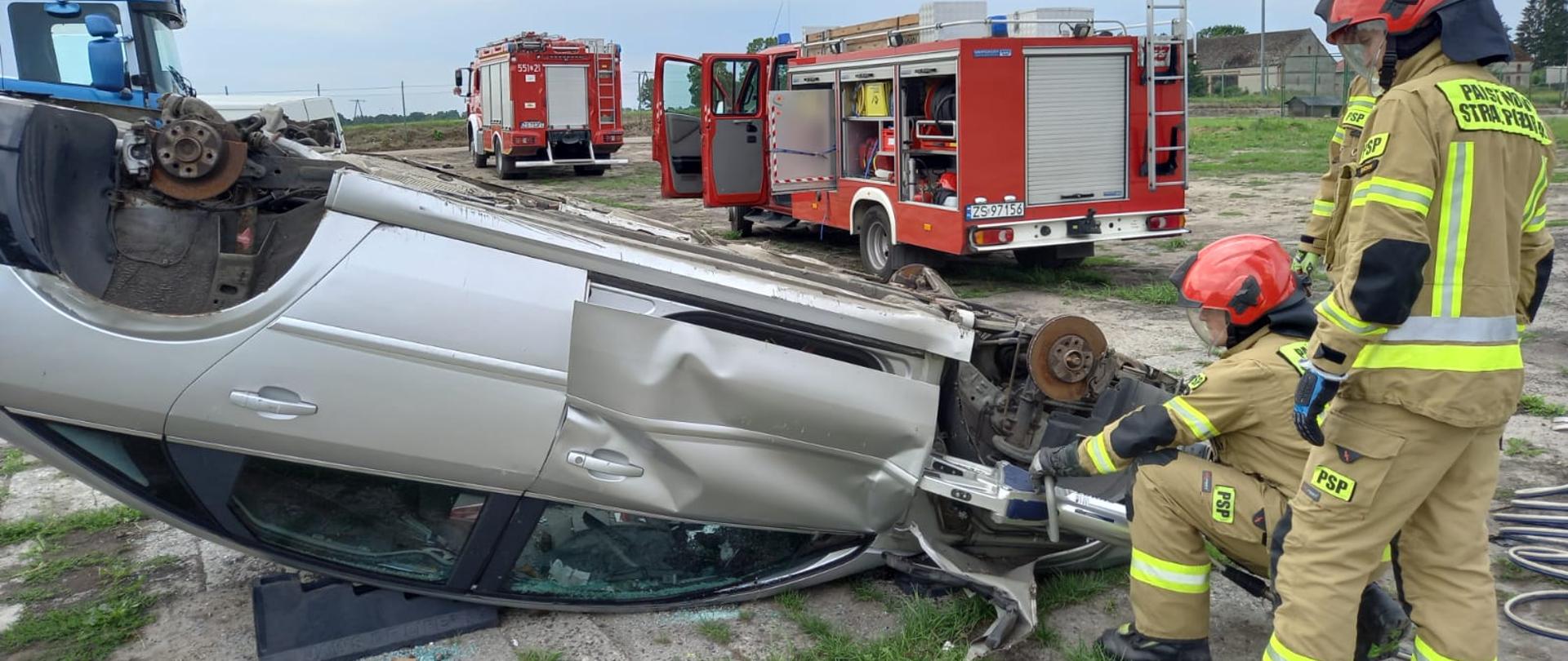 Zdjęcie przedstawia wrak samochodu leżący na dachu przy którym strażacy wyrywają drzwi przy pomocy rozpieracza ramieniowego.