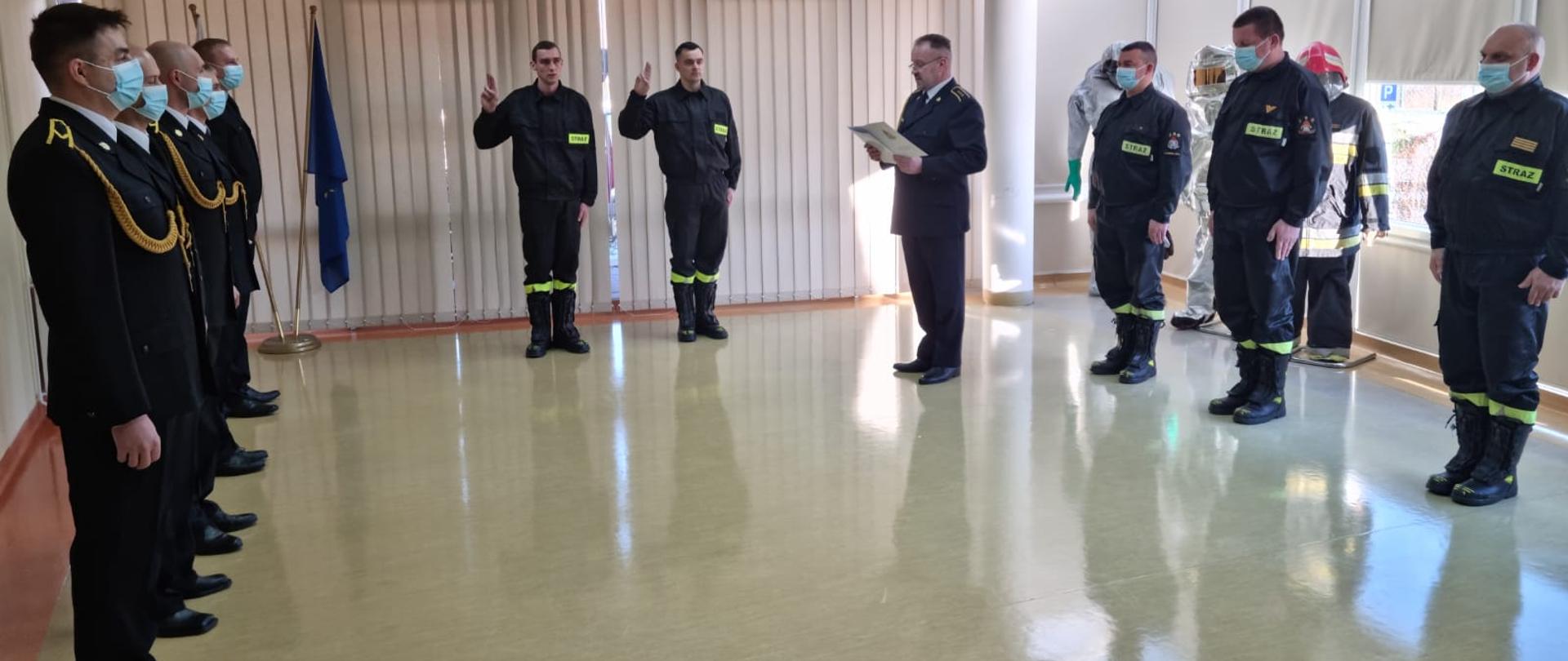 Sześciu funkcjonariuszów ustawionych po lewej stronie w postawie zasadniczej otrzymujących wyróżnienia za zaangażowanie w działaniach poza granicami Kraju. Uroczystość odbywa się w sali sztabowej w tle na ścianie widoczne Godło Polski. Po prawej stronie dostrzegamy Zastępcę Jednostki Ratowniczo Gaśniczej czytającego słowa przysięgi. Po prawej stronie zdjęcia ustawieni trzej funkcjonariusze w ubraniach służbowych.