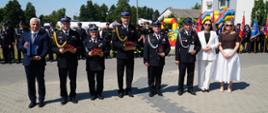 Na zdjęciu widać grupę osób stojących w szeregu podczas uroczystości strażackiej. Pięciu mężczyzn w mundurach strażackich oraz jeden mężczyzna w garniturze trzymają w rękach statuetki i tablice pamiątkowe. Obok nich stoją dwie kobiety – jedna w białym garniturze, druga w brązowej bluzce i białej spódnicy. W tle znajdują się strażacy w mundurach, poczty sztandarowe oraz kolorowy dmuchaniec, co wskazuje na charakter święta lokalnej społeczności. Całość odbywa się na świeżym powietrzu, w słoneczny dzień.