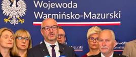 Wojewoda warmińsko-mazurski Radosław Król przemawia podczas briefingu prasowego. W tle widoczna jest ścianka Wojewody Warmińsko-Mazurskiego oraz grupa oficjeli uczestniczących w wydarzeniu.