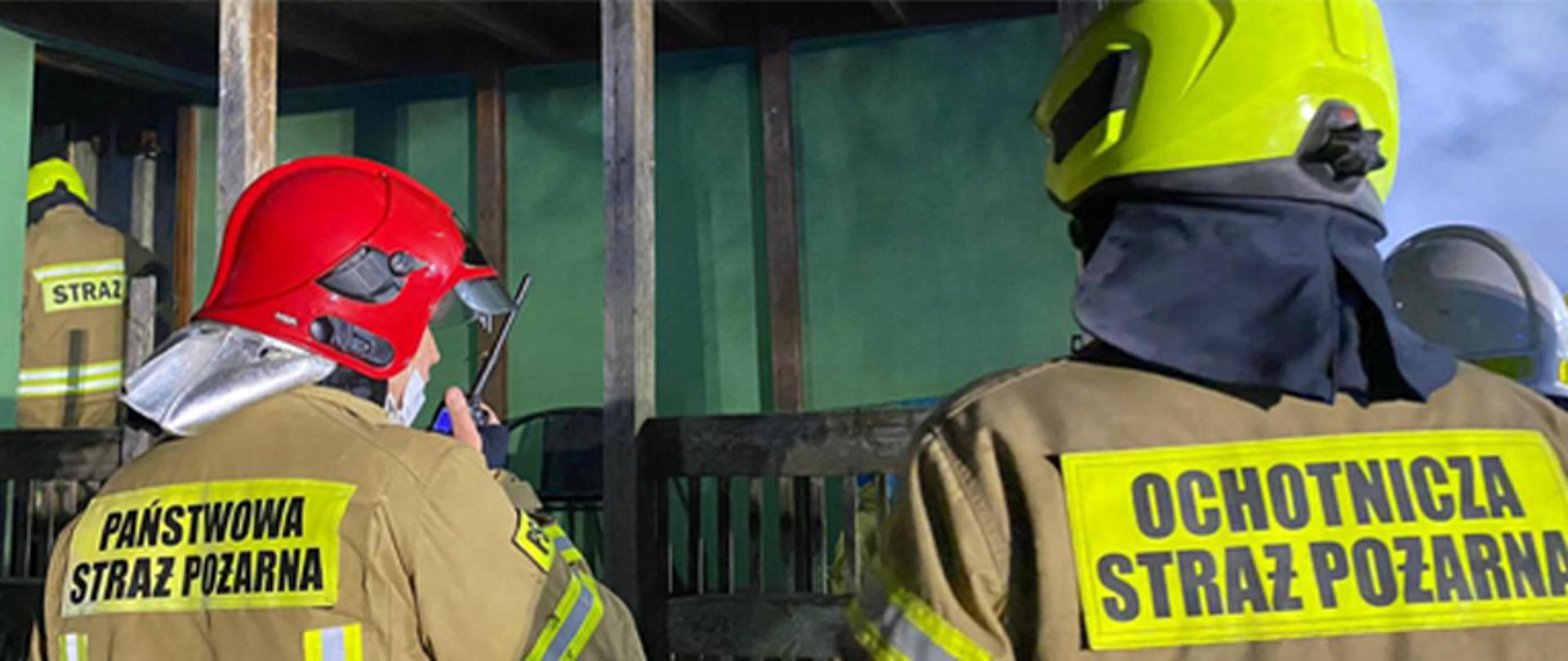 Dwóch strażaków odwróconych plecami do zdjęcia, ubrani są w umundurowanie bojowe typu Moratex barwy jasnobrązowej, strażak po lewej stronie kadru ma na głowie hełm barwy czerwonej oraz napis na plecach PAŃSTWOWA STRAŻ POŻARNA i rozmawia przez radiostacje przenośną, strażak po prawo ma na głowie hełm barwy żółtej i napis na plecach OCHOTNICZA STRAŻ POŻARNA, w tle po lewej stronie stoi w drzwiach strażak w ubraniu bojowym typu Moratex barwy jasnobrązowej, na głowie hełm barwy żółtej oraz napis na plecach STRAŻ, dna drugim planie wda słupy drewniane, które tworzą balustradę z drewna z pionowymi sztachetami