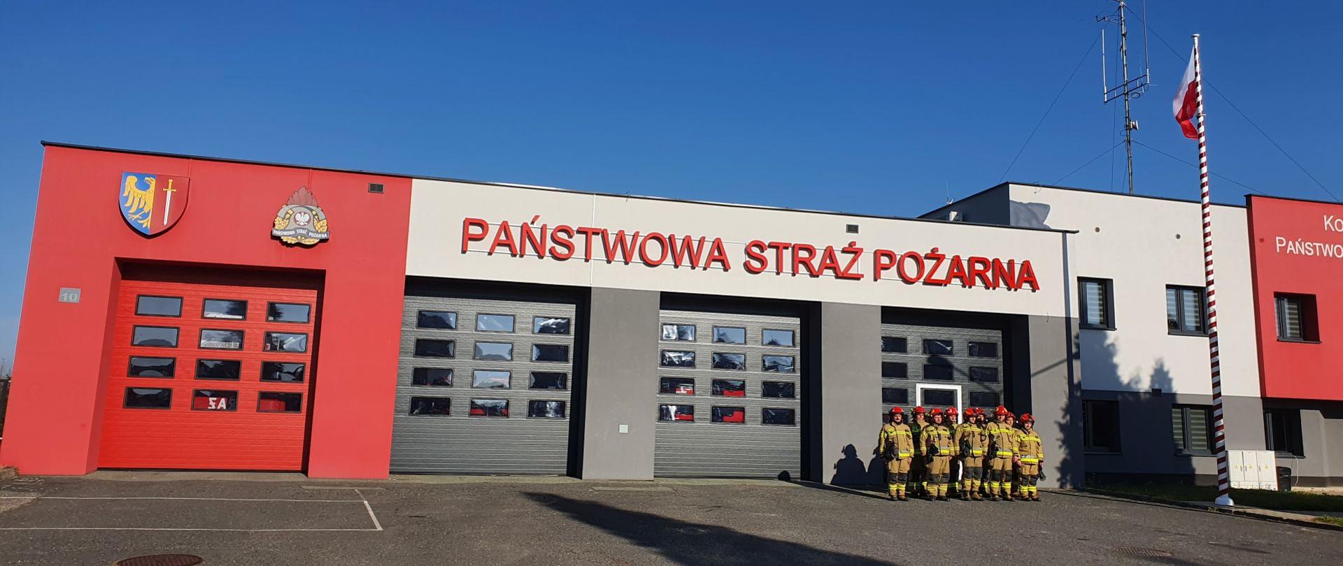 Strażacy w mundurach bojowych stoją w dwuszeregu na tle szarej ściany komendy. Po ich lewej stronie powiewa wciągnięta na maszt flaga państwowa.