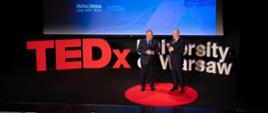 Wiceminister Gzik i mężczyzna w garniturze stoją obok siebie na podwyższeniu z przodu zaciemnionej sali, trzymają w rękach mikrofony, za nimi na wielkim ekranie napis TEDx University of Warsaw, poniżej pod ekranem napis TEDx University of Warsaw ułożony z dużych liter.