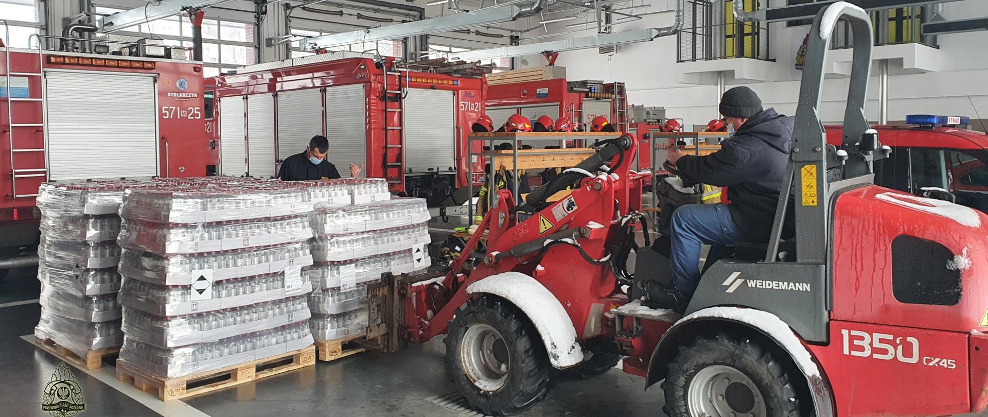 Garaż. Z prawej strony osoba rozładowująca wózkiem widłowym palety na których znajduje się płyn do dezynfekcji. Z tyłu samochody pożarnicze, w kolorze czerwonym. 