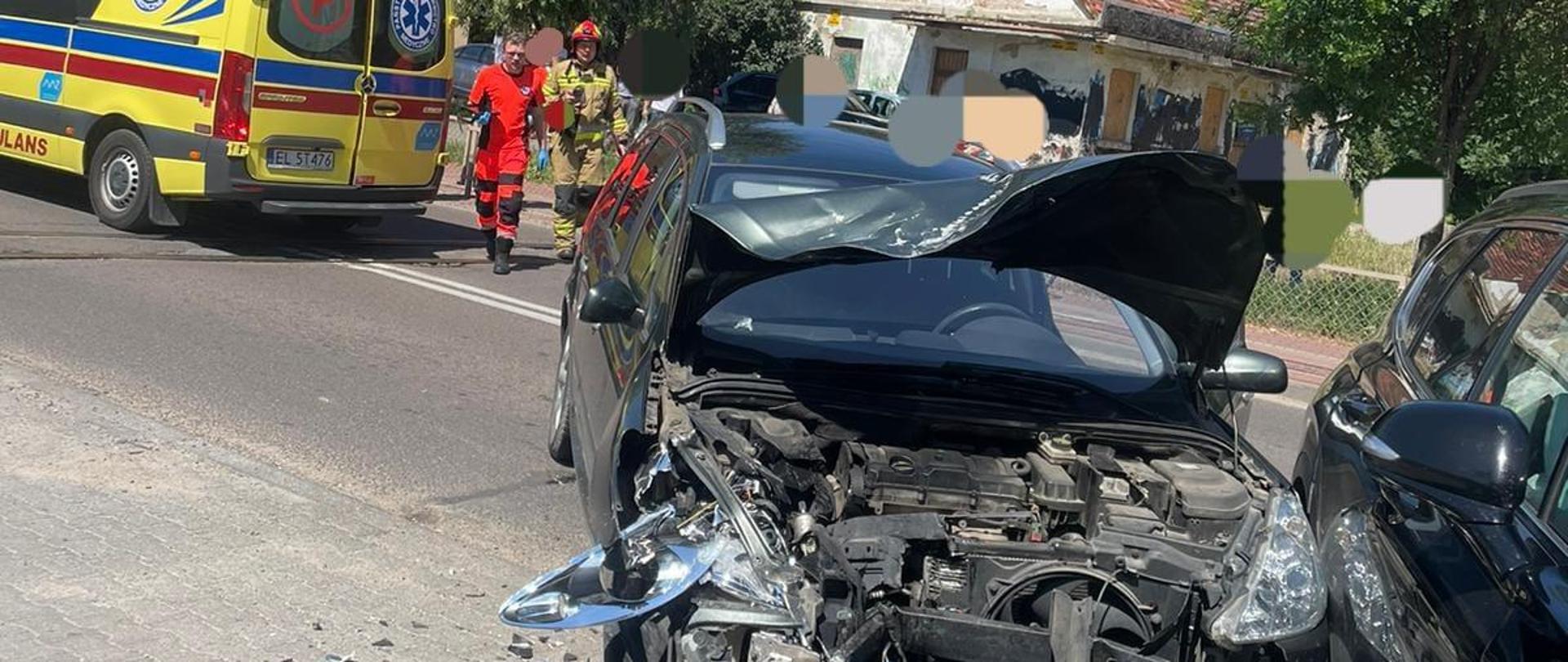 Na zdjęciu widać rozbity ciemny samochód osobowy oraz stojący obok niego drugi ciemny samochód osobowy. Oba pojazdy stoją na asfaltowej jezdni. Na jezdni stoi żółta karetka pogotowia ratunkowego, ratownik medyczny w pomarańczowym stroju oraz strażak w żółtym ubraniu i czerwonym hełmie. W tle widać stary budynek oraz drzewa. Jest widno.
