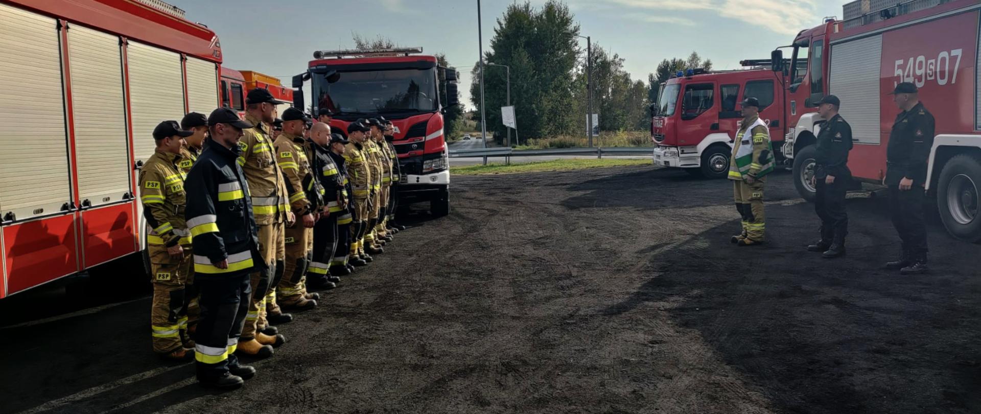 Zdjęcie przedstawia grupę strażaków stojących w dwuszeregu. Wokół rozstawione są pojazdy ratowniczo - gaśnicze.