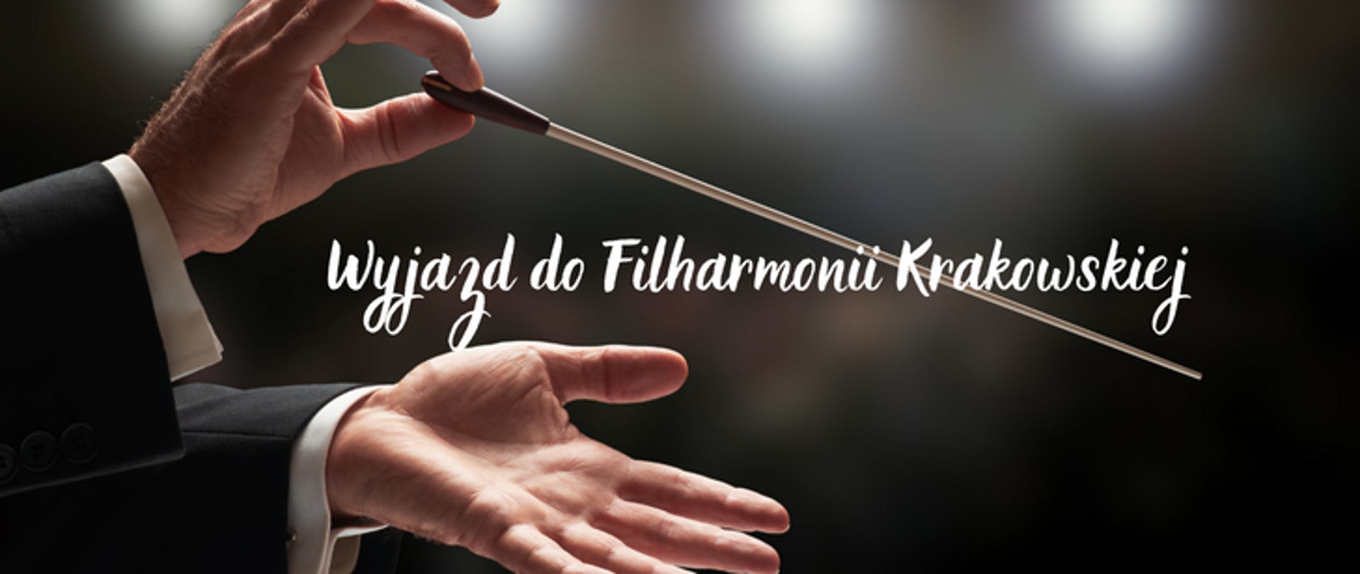 Afisz na ciemnym tle, po lewej stronie ręce dyrygenta trzymającego batutę. Na środku napis Wyjazd do Filharmonii Krakowskiej.