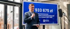 Prezes Wojewódzkiego Funduszu Ochrony Środowiska i Gospodarki Wodnej w Gdańsku przemawia trzymając kartkę. Za nim wyświetlany jest slajd z kwotą 933 675 złotych. 